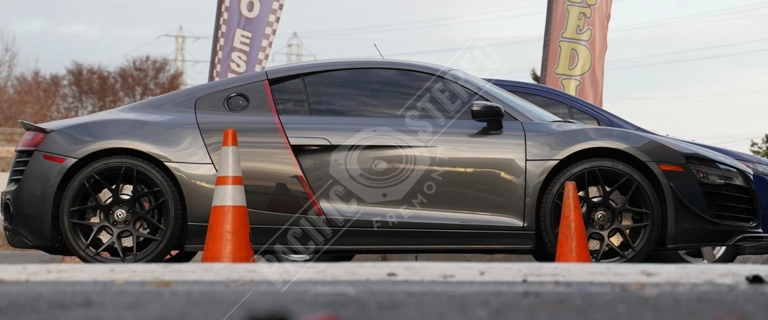 Side view of a sleek grey Audi R8 sports car with black rims and red accent trim, parked between orange traffic cones in a commercial lot. Banners and other vehicles are visible in the background.