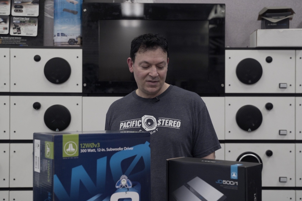 A Pacific Stereo Fremont team member stands in front of car audio equipment displays, reviewing subwoofer and amplifier boxes as part of the process to define your goals & budget for a car audio upgrade.
