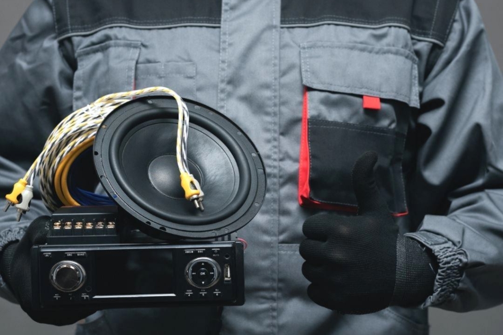 Technician in a work uniform holding a car speaker, stereo head unit, and wiring, representing the process to choose the right aftermarket car audio components.
