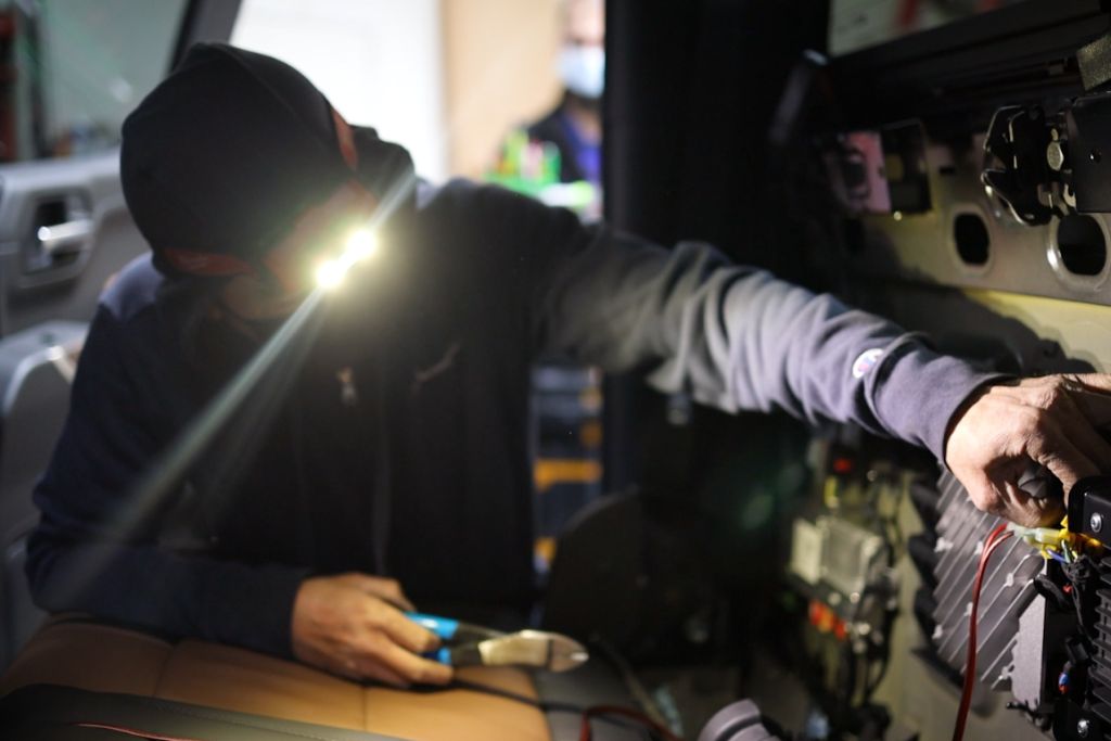 Technician wearing a hoodie and headlamp works inside a vehicle, installing a complex car security system with visible wiring and electronic components. The scene is lit by the headlamp, highlighting the detailed installation process.