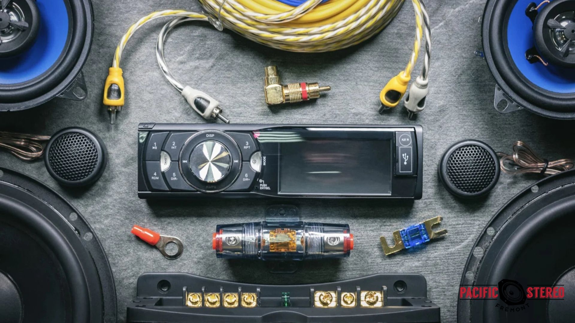 Flat lay of Car Audio equipment including a single din head unit, two coaxial speakers, tweeters, wiring kits, RCA cables, fuse holders, and connectors arranged on a gray surface.