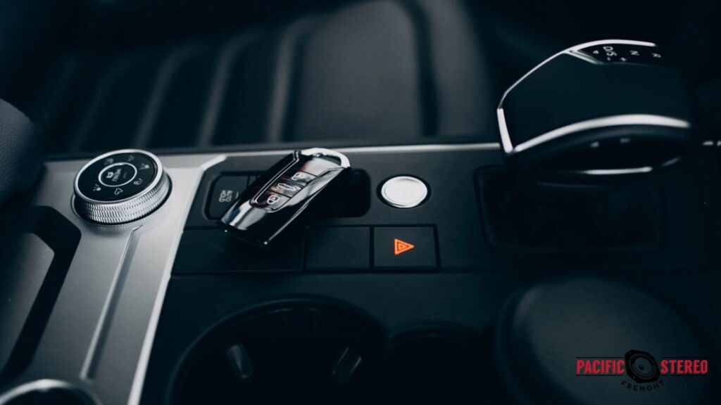 Close up of a vehicle center console with a key fob resting beside the gear selector and illuminated hazard button. The image represents Remote Start convenience by highlighting modern push button ignition and keyless entry features inside the vehicle.
