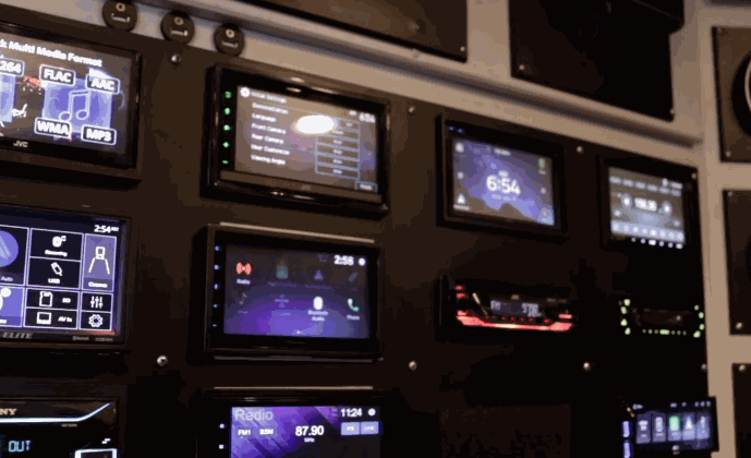 Display wall featuring multiple Car Audio head units with illuminated touchscreens showing radio stations, music apps, and system settings.