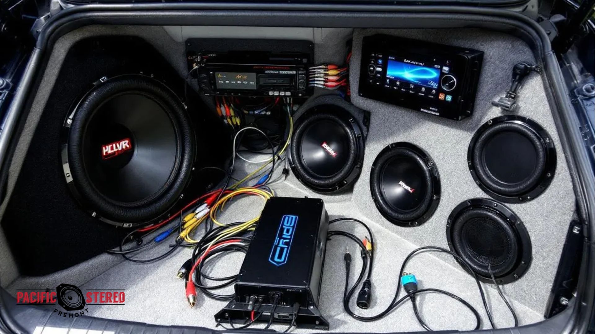 Custom car audio setup installed in a trunk with multiple subwoofers and speakers mounted in a gray enclosure. An amplifier and wiring are visible alongside a digital head unit display showing controls for the car audio system.