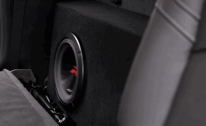 Close up of a subwoofer mounted inside a carpeted enclosure in the rear cargo area of a vehicle as part of a car audio system installation. The speaker cone and enclosure show a compact setup designed to enhance bass in the car audio system.