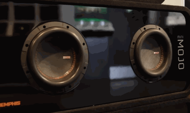 Close up of a Memphis car audio subwoofer enclosure with two round speakers labeled "MOJO mini MOJO" on a glossy black box. The setup highlights a compact dual subwoofer design for enhanced bass inside a vehicle.