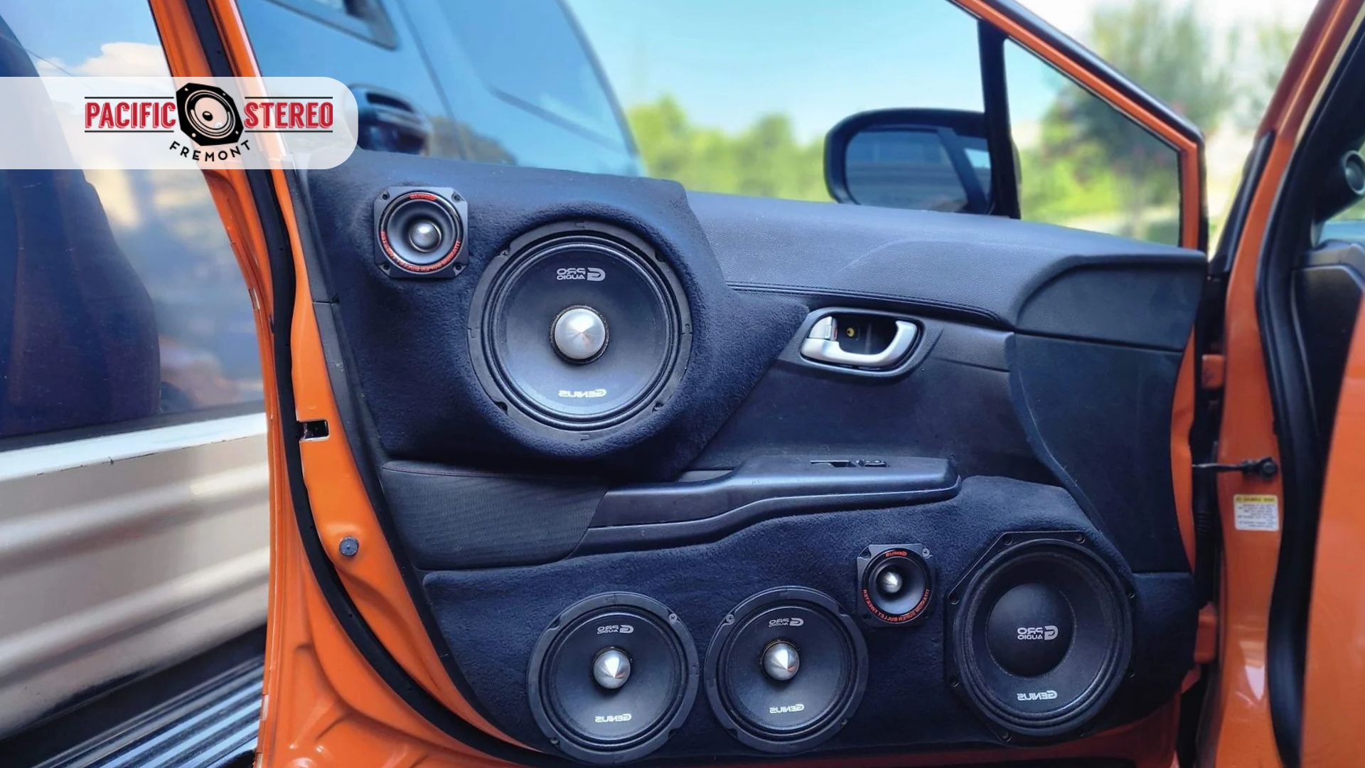 Orange car door opened to show a custom car audio system with multiple black speakers and tweeters built into the door panel. A Pacific Stereo Fremont logo sits in the upper left corner.